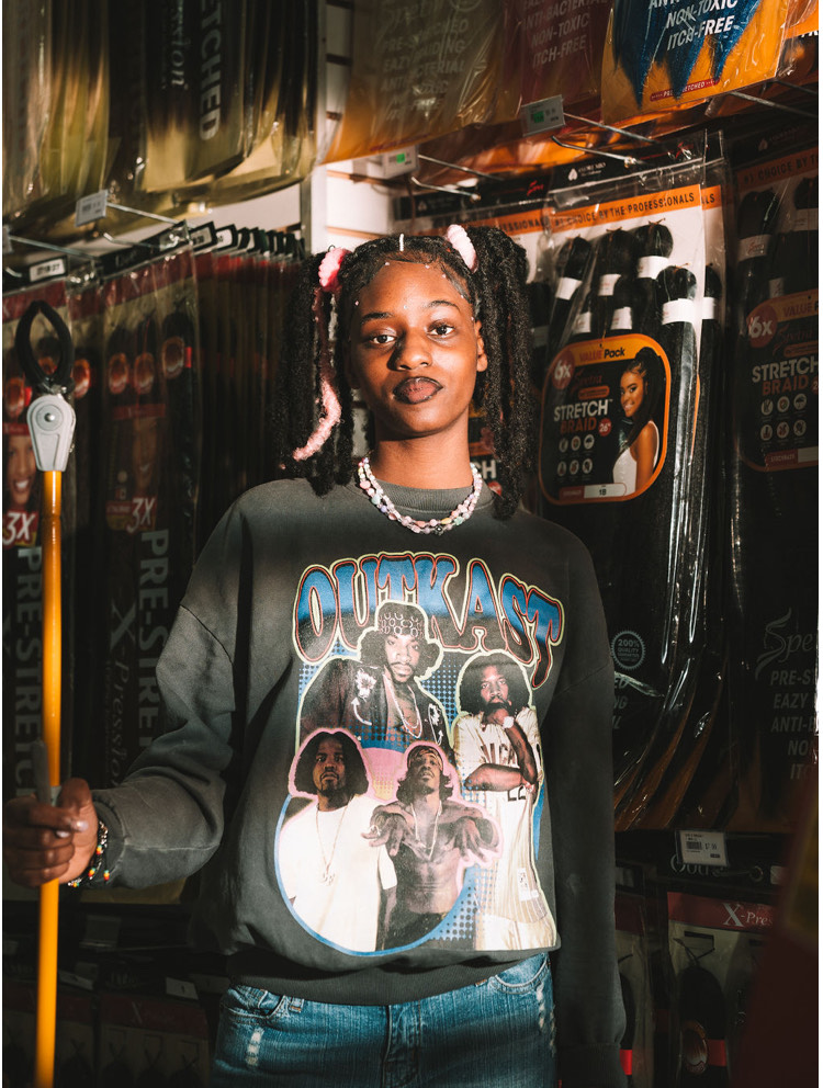 Divinity Nix-Sow stands tall in her local beauty supply store. She wears an Outkast sweatshirt and her hair in pigtails. She holds a tall pole used to grab materials from higher shelves. 