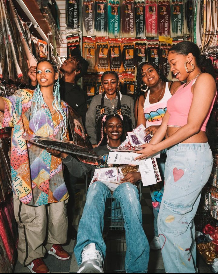 Divinity Nix-Sow sits in a shopping cart piled high with weaves and hair accessories. She is surrounded by her models who smile as they engage with one another. 