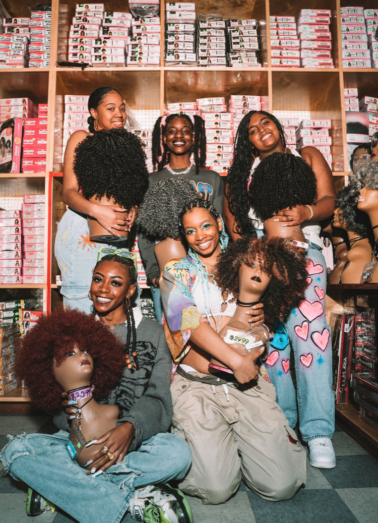 Divinity Nix-Sow poses with four models from her Black History Month natural hair shoot. Each model holds a cosmetology mannequin head, each adorned with a different curly-haired wig. 