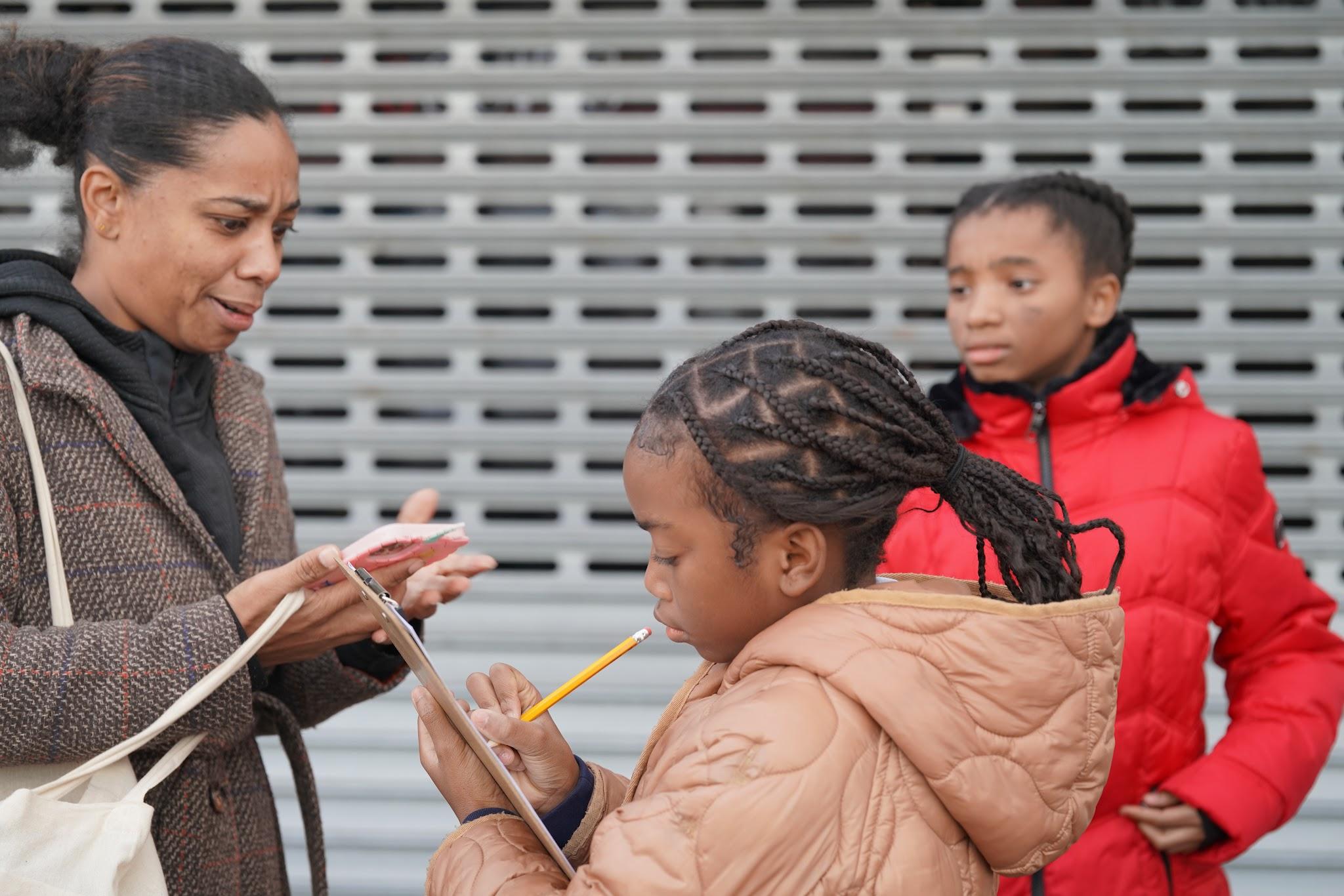 Two elementary-aged students. One is holding a pencil and clipboard. Both are talking with an adult as they conduct community street audits. 