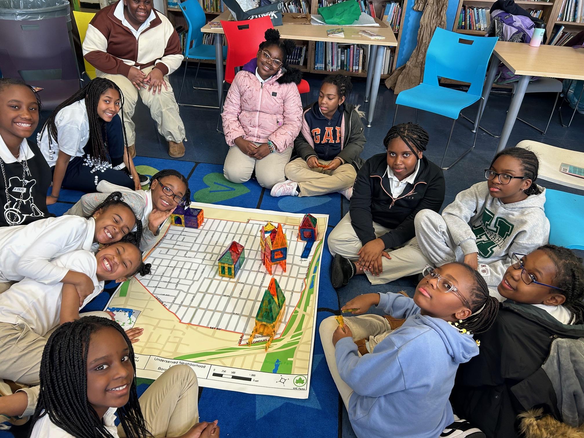 A group of students sitting in a circle around a large floor map and model houses made from Magna-tiles. 