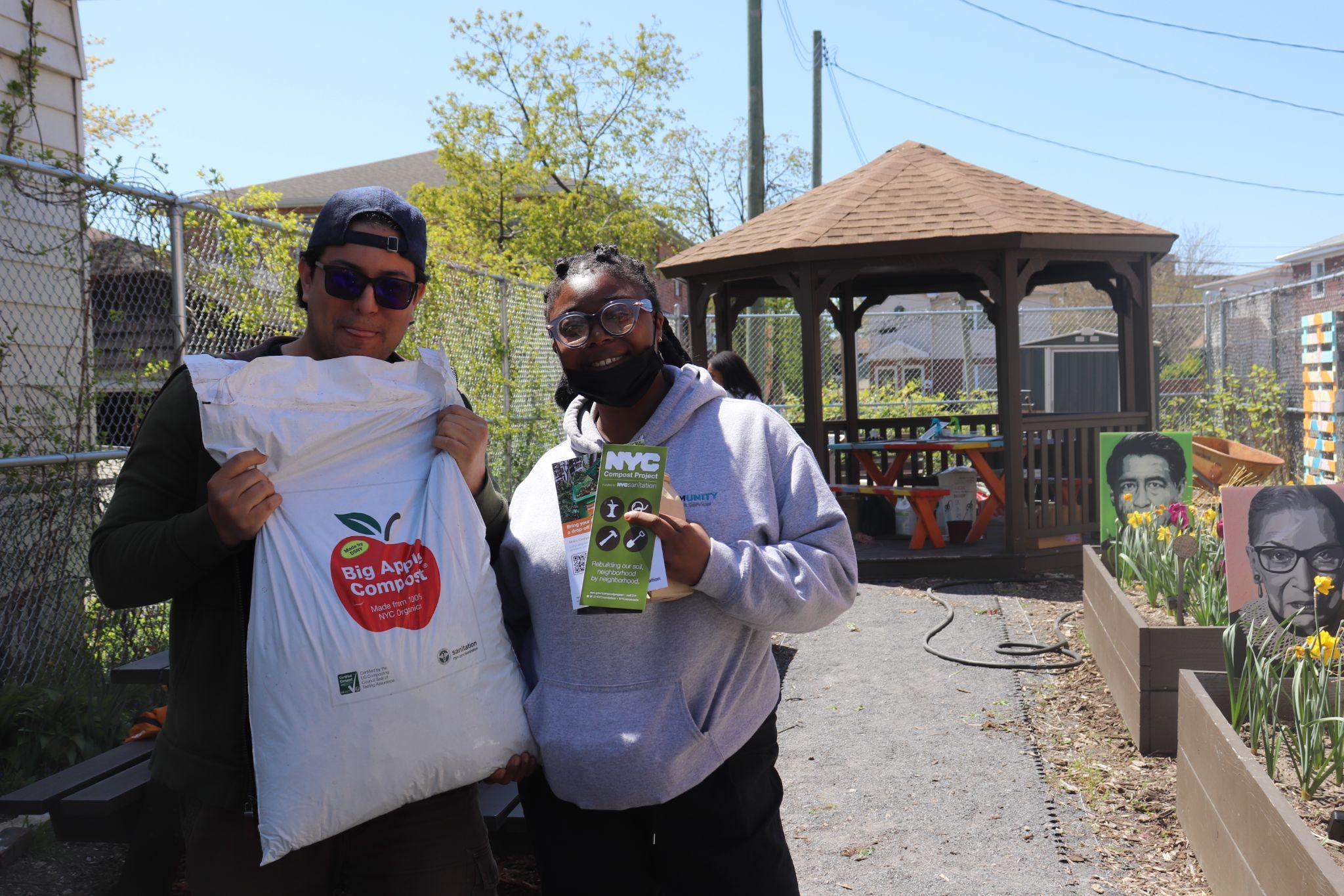 Two community members holding a large bag of compost and standing together in the community garden. 